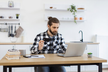Happy Caucasian gentleman doing online banking transaction while working remotely. Attractive bearded man in checkered shirt holding credit card while typing on portable computer in kitchen interior.