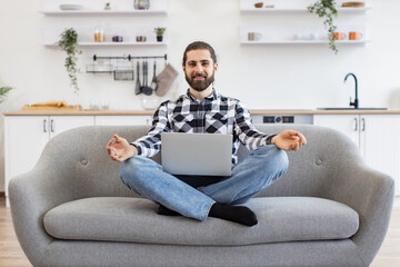 Peaceful Caucasian businessman in casual attire sitting on couch with wireless laptop on knees and meditating. Bearded young guy with fingers in mudra gesture relieve stress and regaining energy. - obrazy, fototapety, plakaty