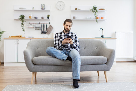 Caucasian male in checkered shirt holding smartphone while staying in modern workplace at home. Efficient freelance worker texting message to colleague while performing project for employer indoors.