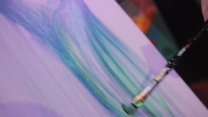 Close up view of talented artist hand holding paintbrush and applying strokes of paint to canvas. Beautiful masterpiece adorned with abstract and flowing lines of blue and green hues.