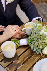 Hands of the groom and the bride, bouquet of flowers, and coffee cups on the table