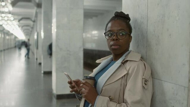 Side chest up footage of 30 yo stylish Black lady in glasses waiting for her train at subway station while checking messages on phone and holding disposable coffee cup