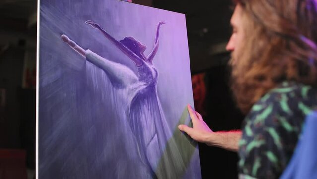 View from shoulder of fine artist sitting near easel painting indoors. Elegant artwork portraying ballet dancer wearing flowing dress frozen in mid-motion with gracefully extended arms.
