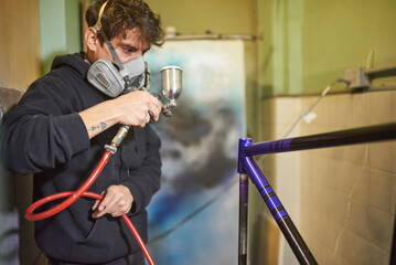 Latin young man wearing a respirator mask concentrated spray painting a bicycle frame in his...