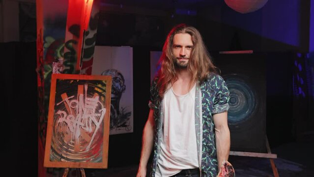Ingenious artist dressed in green patterned open shirt standing next to easel in creative art space. Talented caucasian male with long hair presenting work at exhibition indoors.