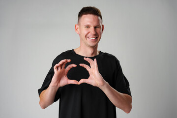Smiling young man gesturing heart sign with ahnds against gray background
