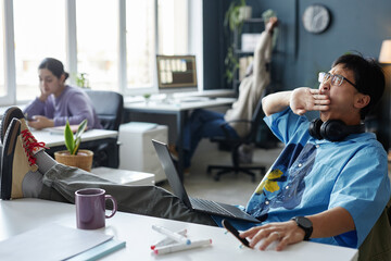 Side view portrait of Asian young man yawning while working with laptop in office feet up on table copy space