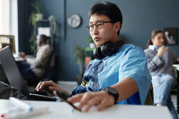 Portrait of creative Asian man with headphones using laptop at office workplace copy space - obrazy, fototapety, plakaty