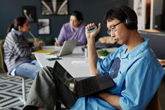 Side view portrait of young Asian man looking at laptop screen in office while casually sitting at workplace with hand expander copy space