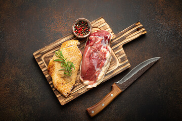Two raw uncooked duck breast fillets with skin, seasoned with salt, pepper, rosemary top view on wooden cutting board with knife, dark brown concrete rustic background. - 761507257