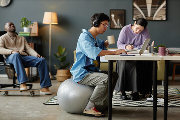Side view full length portrait of Asian man working in office and sitting on fitness ball copy space