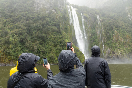 Milford Sound, Fiordland, New Zealand _ Nov. 14, 2023: Tourists snap pictures of a magnificent waterfall froim a ferry on Milford Sound, one of the wettest places on the planet.