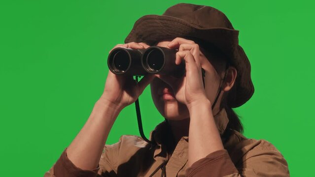 Young female tourist in khaki and hat looking through binoculars during hike in nature while standing on green chroma key background