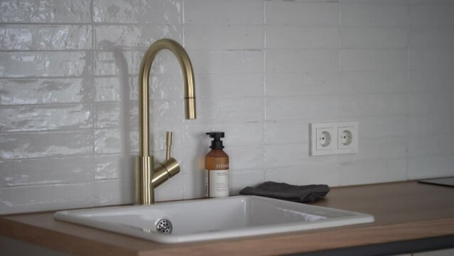 Cesis, Latvia - February 14, 2024 -  a kitchen sink with a modern gold faucet, beside a soap dispenser and a cloth, set against a tiled backsplash with electrical outlets.
