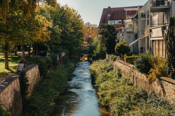 Fototapeta premium Beautiful small river with clean and clear water front of colorful autumn trees and small old town on the hill agaist nice blue and clouds sky during autumn in Europe