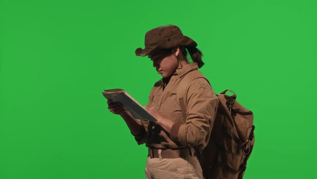 Young female hiker with backpack using orienteering map and looking into distance while standing in studio with green chroma key wall
