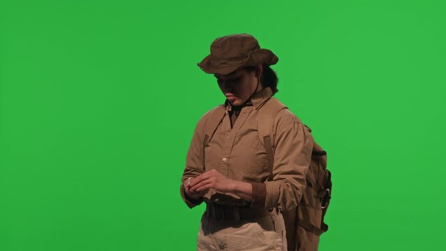 Young female tourist with backpack establishing direction with compass during hike while standing against green chroma key background