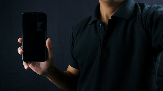 Asian man holding big smartphone with blank screen in hand, showing close to camera and pointing at device. Gadget with empty free space for mock up, banner