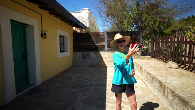Mature woman tourist wearing straw hat and ethnic, traditional clothes taking selfie photo with phone in front of old building in mining town of El Triunfo, Mexico.