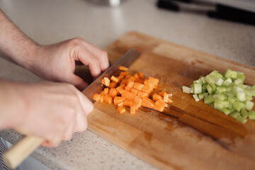 A detailed shot of hands cutting fresh carrots and celery on a wooden board. - obrazy, fototapety, plakaty