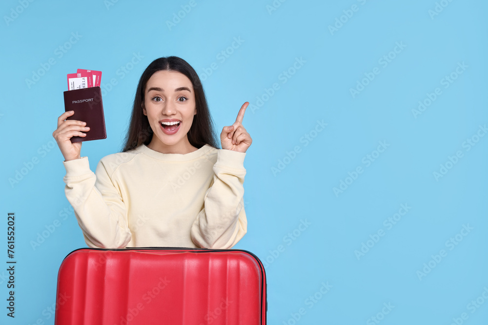 Poster Happy woman with passport, tickets and suitcase pointing at something on light blue background. Space for text