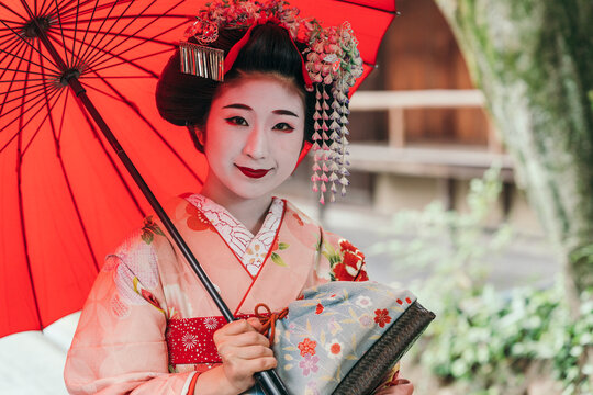 The serene smile of a geisha under her red parasol captures the gentle spirit of traditional Japanese artistry. She exudes cultural beauty and poise.