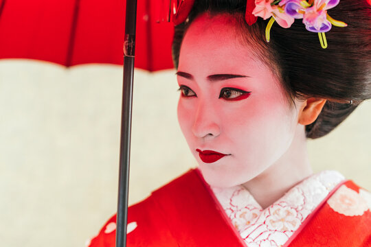 A geisha's intense gaze beneath a red parasol is striking, her red and white kimono embodying the essence of Japanese elegance and tradition
