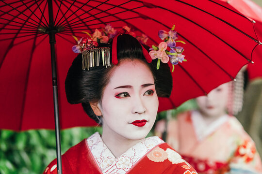 A geisha's reflective gaze under a red umbrella captures the depth of Japanese cultural heritage. Her red kimono is a visual symphony of tradition and craftsmanship