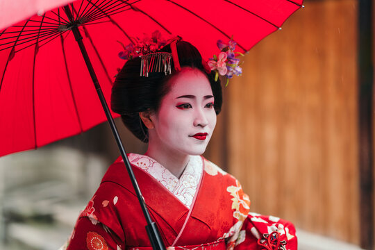 Close-up of a geisha in a red kimono, her expression serene against the backdrop of her red parasol. The image reflects the detailed beauty of geisha makeup and attire