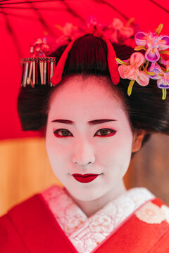 A geisha’s face is perfectly framed by her red parasol, highlighting her striking makeup and the fine details of her hair ornaments. The image exudes traditional Japanese elegance