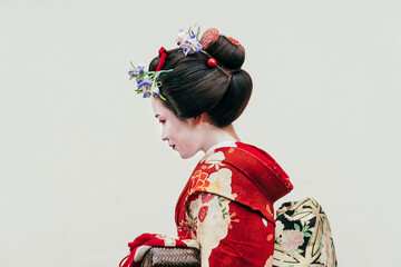 A maiko in a traditional red kimono looks down, showcasing her elaborate hairstyle adorned with floral accessories against a pale backdrop