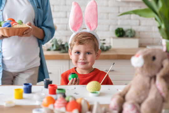 Happy small boy in bunny ears painting eggs for Easter with his pregnant mom in the background. Family celebration. Little kid child son playing with mother while decorating for holiday at home