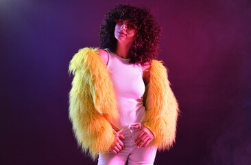Beautiful young woman in yellow fur coat and sunglasses on color background in neon lights