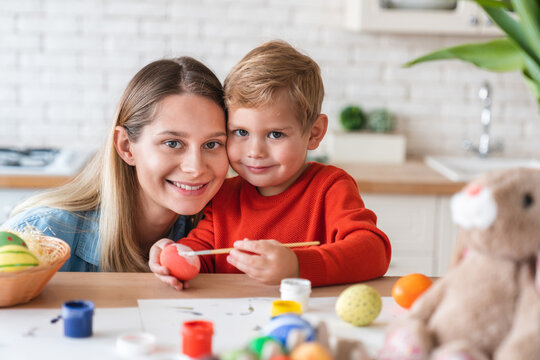 Cheerful caucasian mother and her little son painting eggs for Easter at home. Portrait of happy mom and kid child boy looking at camera while decorating for spring festive celebration