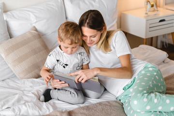 Loving caring young mother reading a book with her little toddler son lying on a bed together. Weekend pastime with family. Stay at home, children care concept