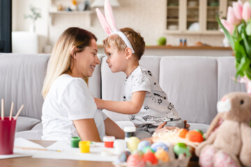 Obraz na płótnie Canvas Cheerful mom and her little son playing together while painting eggs for Easter at home. Young family of two, single mother spending holiday time with child kid