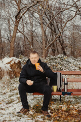 Fototapeta premium Winter Serenity: 40-Year-Old Man Enjoying Tea on Snow-Covered Bench in Rural Park. Immerse yourself in the tranquil beauty of winter as a 40-year-old man finds solace on a snowy bench in a rural park.