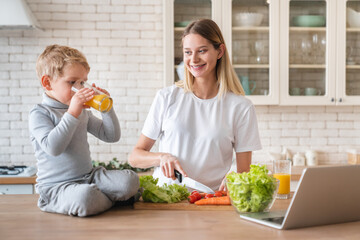 Cheerful little small kid son drinking orange juice in the kitchen helping his mother in cooking vegetable salad in the kitchen together. Family time, healthy eating concept