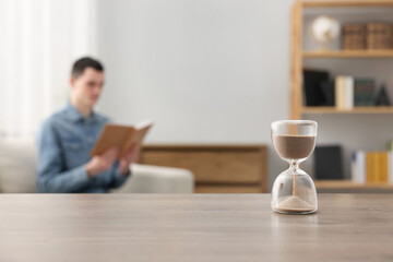 Hourglass with flowing sand on desk. Man reading book in room, selective focus