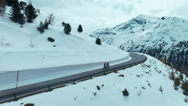 Cinematic drone shot, camera follows two cyclists ride uphill on the way to mountain top, snow covered road in early spring in Alps. Professional training camp before start of season