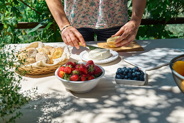 Anonymous woman making sweet mini sandwiches for healthy breakfast in the garden. Toast with cream cheese, blueberry, strawberry, coconut flakes and herbs for summer picnic or brunch