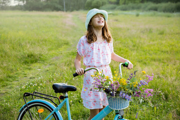 Girl on a bike with a basket of wild flowers on a meadow. Sunny summer day, rays of light, rural background. Summer vacation concept.