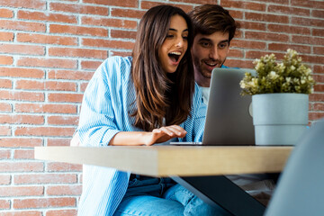 Excited couple make winner expression in front of an open laptop after win money. Successful moment at home with young man and woman watching computer together. Surprise and happiness online business