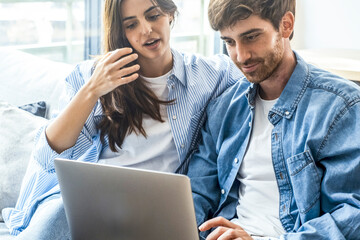 Couple in blue denim casual clothes using together a laptop sitting comfortable on the sofa at home during sunday afternoon. People working on computer at home indoor leisure online activity