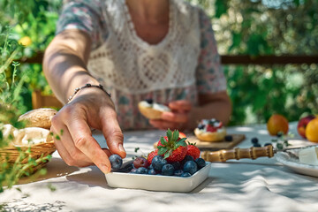 Anonymous woman making sweet mini sandwiches for healthy breakfast in the garden. Toast with cream cheese, blueberry, strawberry, coconut flakes and herbs for summer picnic or brunch