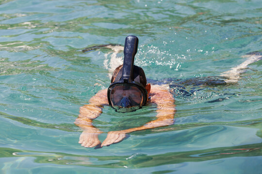 Caucasian man snorkeling in clear waters with black goggles and a snorkel, arms are extended forward as they swim. Male wearing with the  black snorkel full face mask diving above the water in sea