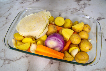 A glass dish filled with vegetables including carrots, potatoes, and onions. The dish is on a marble countertop