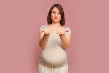 A pregnant woman shows her palms with a plea for help on a studio pink background. Pregnancy in a woman with a belly