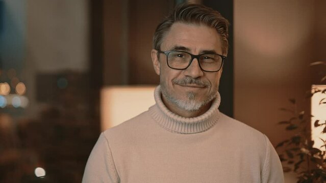 Portrait of confident, elegant middle aged man smiling, standing in living room at home or in hotel apartment. Happy male in glasses and turtleneck pullover.
