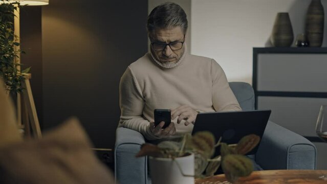 Middle aged man sitting in comfort in living room working on laptop computer and phone in home office. Businessman managing business online. Portrait of confident older male, happy, smiling.